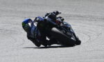 MotoGP rider in a blue racing suit leans into a sharp turn on a race track, riding a Yamaha bike with Monster Energy decals visible.
