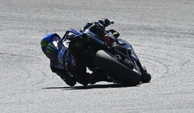 MotoGP rider in a blue racing suit leans into a sharp turn on a race track, riding a Yamaha bike with Monster Energy decals visible.
