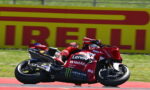 MotoGP rider on a red Ducati racing bike leaning into a corner on a race track, with sponsor logos like Lenovo and Monster Energy visible on the bike.