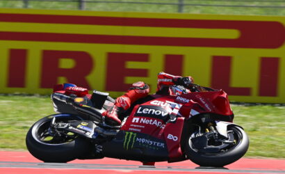 MotoGP rider on a red Ducati racing bike leaning into a corner on a race track, with sponsor logos like Lenovo and Monster Energy visible on the bike.