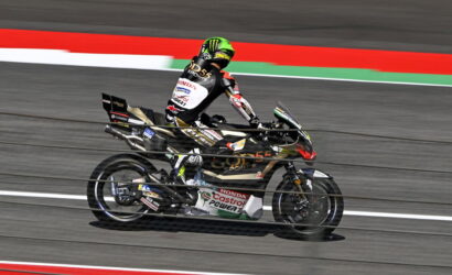 Motorcycle racer in a black-gold Honda livery speeds along a race track, neon green helmet visible.