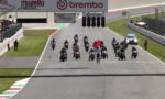 Motorcycle racers line up at the start on a race track as a marshal waves a red flag in front, banners overhead show Mugello Circuit and Brembo logos.