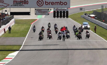 Motorcycle racers line up at the start on a race track as a marshal waves a red flag in front, banners overhead show Mugello Circuit and Brembo logos.