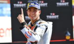 Smiling motorcycle racer in a white suit with sponsor logos, raising one finger in celebration in front of a Tissot backdrop.
