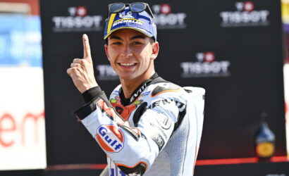 Smiling motorcycle racer in a white suit with sponsor logos, raising one finger in celebration in front of a Tissot backdrop.