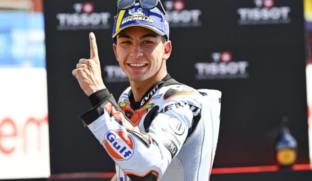 Smiling motorcycle racer in a white suit with sponsor logos, raising one finger in celebration in front of a Tissot backdrop.
