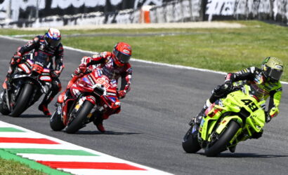 Three motorcycle racers lean into a curve on a race track, with a neon-yellow bike leading the pack behind painted curbs.