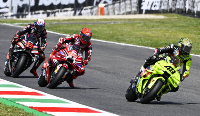 Three motorcycle racers lean into a curve on a race track, with a neon-yellow bike leading the pack behind painted curbs.
