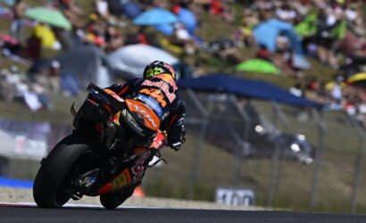 MotoGP rider in Red Bull livery leaning into a turn on a race track, with blurred spectators in the background