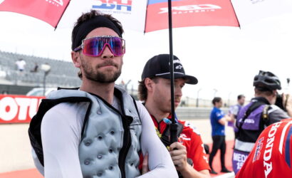Man in a padded racing vest and white sleeves stands with arms crossed under a red umbrella, wearing bright pink mirrored sunglasses and a black headband.