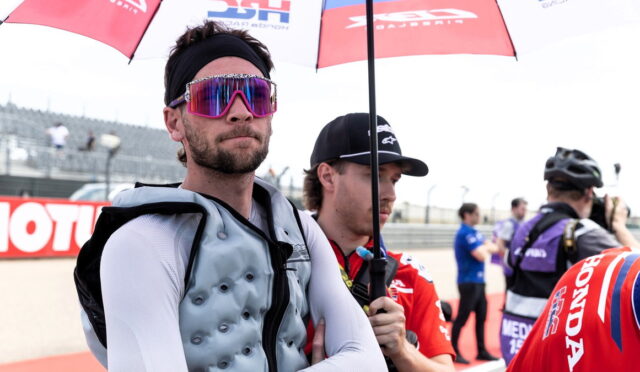 Man in a padded racing vest and white sleeves stands with arms crossed under a red umbrella, wearing bright pink mirrored sunglasses and a black headband.