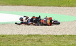 Two motorcycle racers crash-land on a gravel track, sliding side-by-side with bikes skidding and debris around them on the hairpin bend.