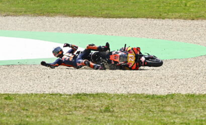Two motorcycle racers crash-land on a gravel track, sliding side-by-side with bikes skidding and debris around them on the hairpin bend.