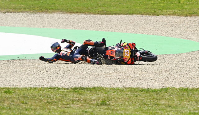 Two motorcycle racers crash-land on a gravel track, sliding side-by-side with bikes skidding and debris around them on the hairpin bend.