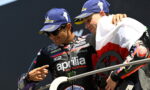 Two Aprilia Racing riders on a podium, one pointing at the other while wearing blue Michelin caps and dark sunglasses.