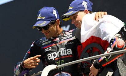 Two Aprilia Racing riders on a podium, one pointing at the other while wearing blue Michelin caps and dark sunglasses.
