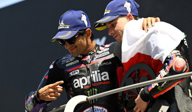 Two Aprilia Racing riders on a podium, one pointing at the other while wearing blue Michelin caps and dark sunglasses.