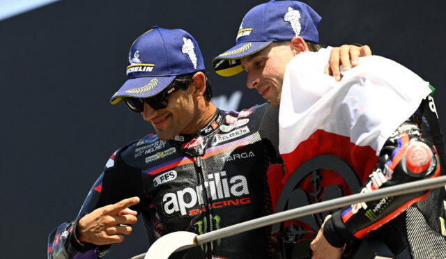 Two MotoGP riders celebrate on a podium, wearing blue Michelin caps and an Aprilia Racing suit with a swaggering gesture.