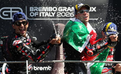 Three motorbike racers celebrate on a podium as champagne sprays around them, with an Italian flag draped over one rider and dark backdrop behind.