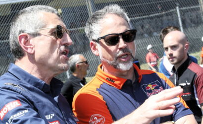 Two men wearing sunglasses and team uniforms talk at a race track, with a chain‑link fence in the background. Alt: Informative caption focusing on conversation at a motorsport event.