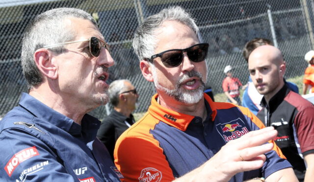Two men wearing sunglasses and team uniforms talk at a race track, with a chain‑link fence in the background. Alt: Informative caption focusing on conversation at a motorsport event.