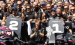 Group of MotoGP riders and crew celebrating at the podium, standing behind large '1' and '2' boards, with cheering fans in the background.