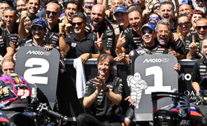 Group of MotoGP riders and crew celebrating at the podium, standing behind large '1' and '2' boards, with cheering fans in the background.