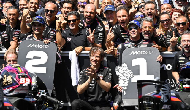 Group of MotoGP riders and crew celebrating at the podium, standing behind large '1' and '2' boards, with cheering fans in the background.
