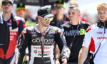 MotoGP rider in black Honda racing leathers with a Monster cap and mirrored sunglasses leads a paddock lineup. Behind him are team members and other riders in racing gear.