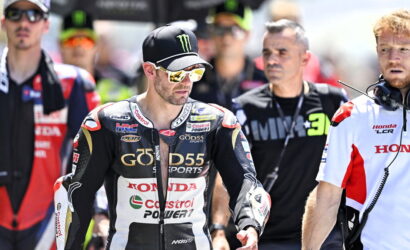 MotoGP rider in black Honda racing leathers with a Monster cap and mirrored sunglasses leads a paddock lineup. Behind him are team members and other riders in racing gear.