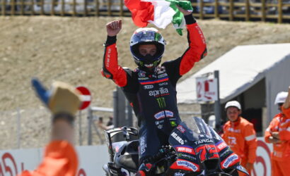 Moto racer in red and black leathers raises both arms, waving an Italian flag from atop a racing bike after winning.