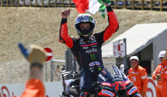 Moto racer in red and black leathers raises both arms, waving an Italian flag from atop a racing bike after winning.