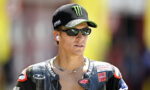 MotoGP rider wearing a Yamaha cap, sunglasses, and wireless earbuds, looking off to the side.