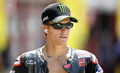 MotoGP rider wearing a Yamaha cap, sunglasses, and wireless earbuds, looking off to the side.