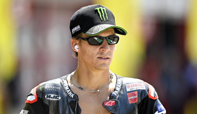 MotoGP rider wearing a Yamaha cap, sunglasses, and wireless earbuds, looking off to the side.