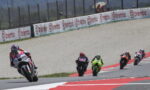 Group of motorcycle racers leaning into a curve on a race track, lead rider on a white bike in red/white gear; sponsor banners along the barrier and red/white curbs visible.