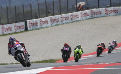 Group of motorcycle racers leaning into a curve on a race track, lead rider on a white bike in red/white gear; sponsor banners along the barrier and red/white curbs visible.