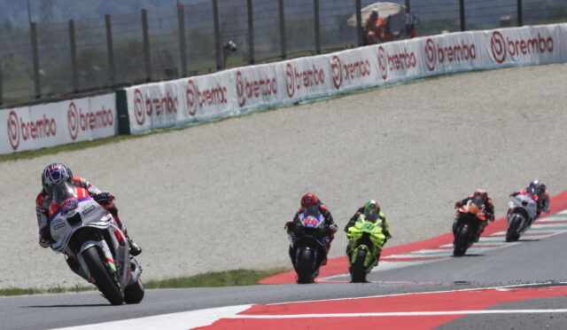 Group of motorcycle racers leaning into a curve on a race track, lead rider on a white bike in red/white gear; sponsor banners along the barrier and red/white curbs visible.