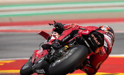 Red Ducati MotoGP rider leans into a sharp turn on a race track, bright red suit and bike with sponsor decals visible on the bike and suit.