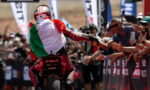 MotoGP rider in red gear high-fiving cheering spectators behind a barrier, wearing an Italian flag cape from the back.