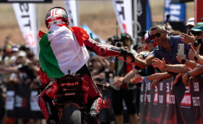 MotoGP rider in red gear high-fiving cheering spectators behind a barrier, wearing an Italian flag cape from the back.