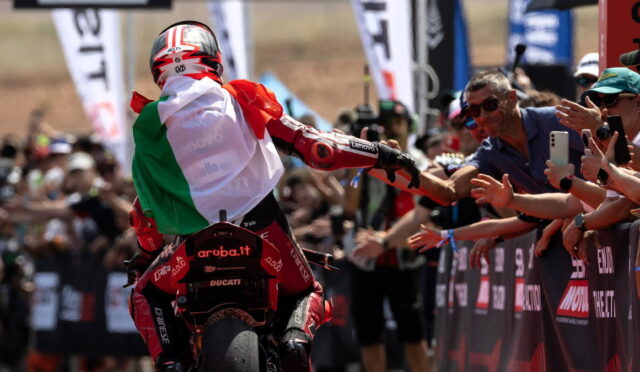 MotoGP rider in red gear high-fiving cheering spectators behind a barrier, wearing an Italian flag cape from the back.