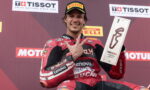 MotoGP rider in red Ducati leathers on a podium, flashing a victory hand gesture while holding a rectangular trophy with a red squiggle logo in front of sponsor banners.