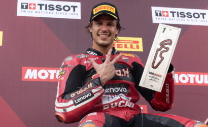MotoGP rider in red Ducati leathers on a podium, flashing a victory hand gesture while holding a rectangular trophy with a red squiggle logo in front of sponsor banners.