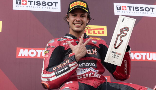MotoGP rider in red Ducati leathers on a podium, flashing a victory hand gesture while holding a rectangular trophy with a red squiggle logo in front of sponsor banners.
