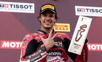 Motocycle racer on a podium in red and black gear, wearing a Pirelli cap, flashing a gesture while holding a silver trophy.