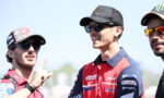 Three motorsport team members in branded caps and sunglasses chat outdoors, with the left man pointing with his finger while smiling at the others.