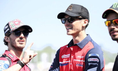 Three motorsport team members in branded caps and sunglasses chat outdoors, with the left man pointing with his finger while smiling at the others.