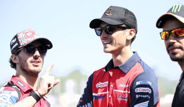 Three motorsport team members in branded caps and sunglasses chat outdoors, with the left man pointing with his finger while smiling at the others.