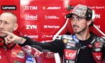 MotoGP rider in a Lenovo-branded racing suit and camo cap points to the left as a team member in red sits beside him in a sponsor-filled red backdrop.